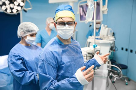 Téléchargez les photos : Le chirurgien est vêtu d'une robe stérile avant l'opération, il est assisté par une infirmière opératrice - en image libre de droit