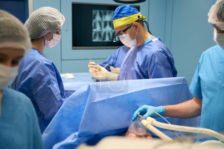Foto de Cirujano con un gorro amarillo-azul toma un instrumento de un asistente, un anestesiólogo le da anestesia a un paciente - Imagen libre de derechos