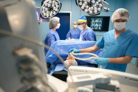 Téléchargez les photos : L'anesthésiste donne la médecine à un patient à travers un masque, une équipe de médecins et d'assistants est à la table chirurgicale - en image libre de droit
