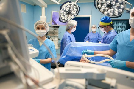 Téléchargez les photos : Anesthésiste, chirurgien, assistants debout à la table d'opération au-dessus du patient, la pièce est éclairée par des lampes opératoires - en image libre de droit