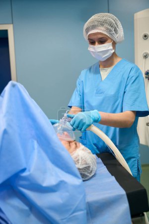 Foto de Anestesióloga mujer da anestesia a la paciente a través de una máscara, un paramédico en un uniforme quirúrgico estéril - Imagen libre de derechos