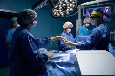 Foto de Ayudante femenina le da al cirujano un instrumento durante la operación, un equipo de médicos trabaja en la mesa quirúrgica - Imagen libre de derechos
