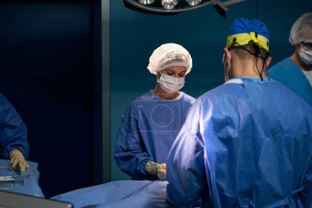 Foto de Asistentes, un anestesiólogo, una enfermera de quirófano y un cirujano trabajan en quirófano, paciente bajo anestesia yace sobre la mesa - Imagen libre de derechos