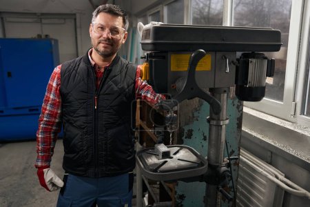 Téléchargez les photos : Homme en vêtements et gants de protection debout près de l'équipement pour percer des détails en acier dans l'atelier - en image libre de droit