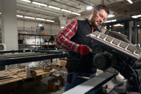Téléchargez les photos : Homme adulte en vêtements et lunettes de protection travaillant sur la machine de découpe dans l'usine - en image libre de droit