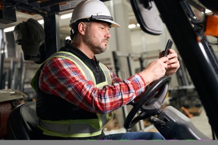 Foto de Hombre profesional en ropa protectora, guantes y casco sentado en la máquina de carga y mirando el teléfono móvil - Imagen libre de derechos