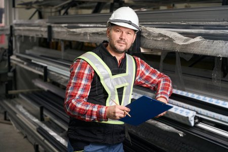 Téléchargez les photos : Travailleur professionnel en vêtements de protection, gants et casque debout près du rack avec des profils métalliques - en image libre de droit