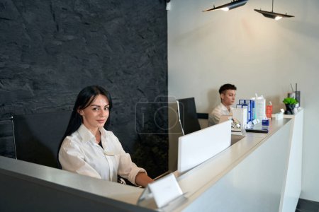 Téléchargez les photos : Jeune femme travaillant à l'ordinateur à côté de son assistant, regardant la caméra, attendant les clients - en image libre de droit