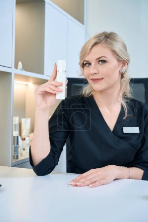 Téléchargez les photos : Portrait de cosmétologue en manteau médical assis dans le bureau, tenant le produit cosmétique et prêt pour le travail - en image libre de droit