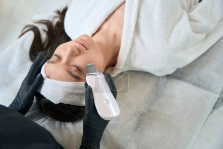Foto de Cosmetólogo en guantes de protección sentado cerca de la mujer adulta y haciendo procedimiento de limpieza facial ultrasónica en el consultorio - Imagen libre de derechos