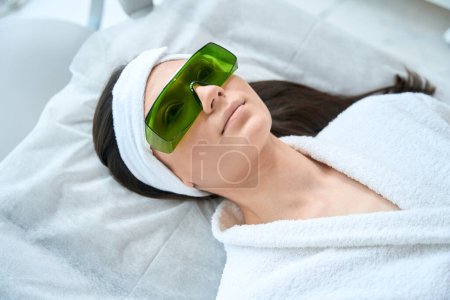 Foto de Mujer joven en gafas protectoras tumbada en el sofá en la oficina del cosmetólogo preparándose para la depilación láser - Imagen libre de derechos
