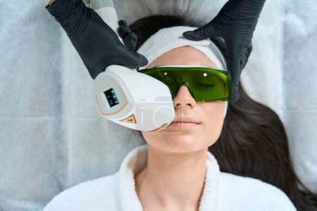 Foto de Enfermera en guantes de protección de pie cerca de la señora adulta y haciendo depilación láser en la cara - Imagen libre de derechos