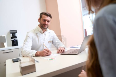 Photo for Waist up portrait of gorgeous bearded doctor is writing the prescription for woman patient in his medical office - Royalty Free Image