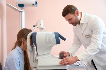 Foto de Cintura hacia arriba retrato de hombre médico está examinando los ojos del paciente con la máquina moderna en el consultorio médico - Imagen libre de derechos