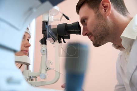 Photo for Side view portrait of handsome Caucasian optometrist is looking through the modern slit lamp while correction the vision of patient - Royalty Free Image