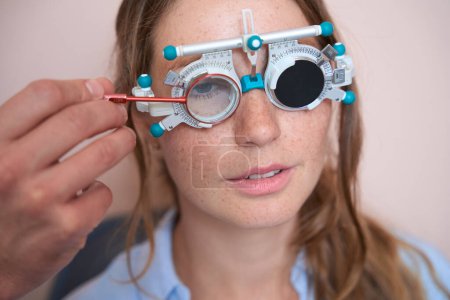 Foto de Primer plano retrato de encantadora mujer elegante está cubriendo un ojo y comprobar su agudeza visual con marco de prueba en la clínica de óptica - Imagen libre de derechos