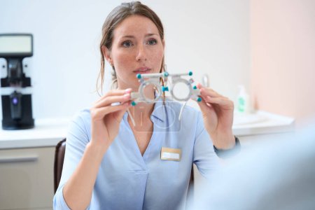 Photo for Waist up portrait of charming Caucasian woman ophthalmologist is holding special professional equipments for the examining eyesight in the medical office - Royalty Free Image