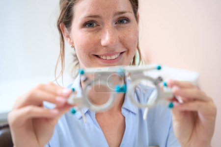 Foto de Retrato de cintura hacia arriba del oftalmólogo femenino caucásico sonriente sostiene equipos profesionales para verificar la agudeza visual - Imagen libre de derechos