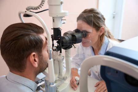 Photo for Side view portrait of smiling doctor optometrist is looking through the slit lamp while examining eyesight of man patient in clinic - Royalty Free Image