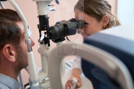 Photo for Close up side view portrait of smiling qualified woman doctor is looking through the slit lamp while examining visual acuity in the medical cabinet - Royalty Free Image