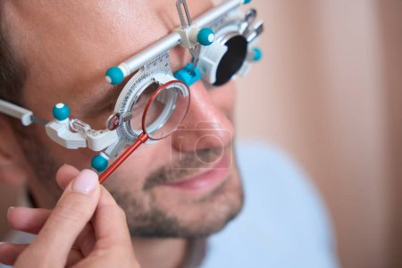 Téléchargez les photos : Portrait latéral d'un beau homme caucasien regarde à travers le cadre d'essai avec des lentilles spéciales dans la clinique d'ophtalmologie - en image libre de droit