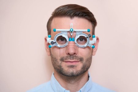 Photo for Close up portrait of handsome Caucasian male is examining his eyesight with trial frame isolated on light pink background - Royalty Free Image