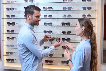 Téléchargez les photos : Portrait de vue latérale de homme barbu et belle femme choisit les nouvelles lunettes de soleil dans le magasin opticien - en image libre de droit