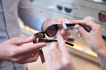 Photo for Cropped head picture of female hands with stylish sunglasses is demonstrating it for the client in optician shop - Royalty Free Image