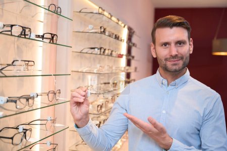 Photo for Close up portrait of bearded Caucasian man is holding the optician frame in front of the shop shelf - Royalty Free Image