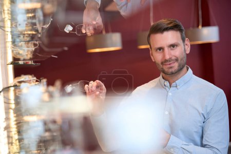 Photo for Waist up portrait if handsome Caucasian male is holding the luxury eye glasses in hand while standing near the shop shelf - Royalty Free Image