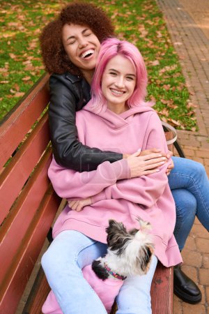 Téléchargez les photos : Agréable jeune femme avec Yorkshire Terrier assis sur le banc dans les bras de sa petite amie joyeuse - en image libre de droit