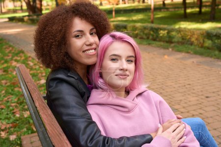 Téléchargez les photos : Heureuse femme caucasienne assise sur le banc dans les bras de sa petite amie afro-américaine aimante - en image libre de droit