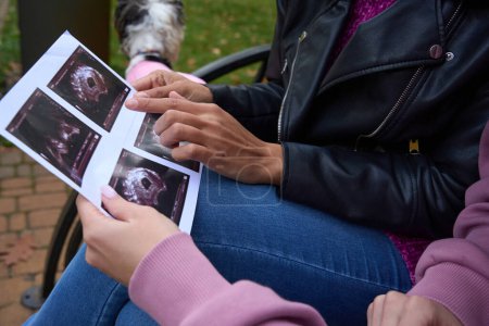 Téléchargez les photos : Photo recadrée d'une dame pointant le fœtus sur une échographie vers sa compagne assise sur un banc de parc - en image libre de droit