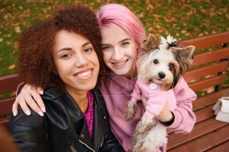 Foto de Feliz pareja de lesbianas de raza mixta tomando selfie con su Yorkshire Terrier en el banco del parque - Imagen libre de derechos