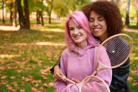 Photo for Waist-up portrait of joyful young woman with badminton racket in hand hugging her pleased female companion outdoors - Royalty Free Image