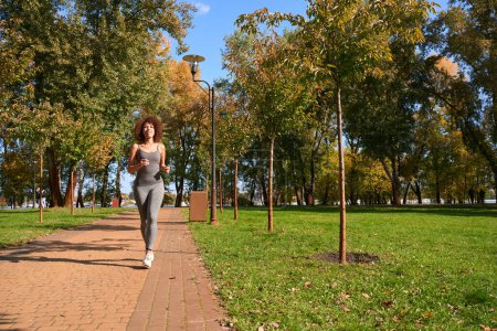 Photo for Full-size portrait of smiling fit young woman running along paved footpath in park - Royalty Free Image