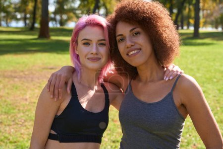 Photo for Waist-up portrait of two joyous athletic women embracing one another in park - Royalty Free Image