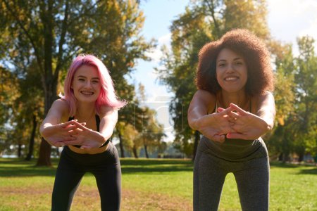 Photo for Two smiling cheerful fit young women performing forward bends in public park - Royalty Free Image