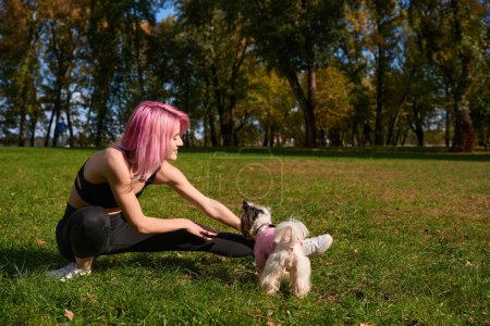 Téléchargez les photos : Agréable jeune athlète féminine faisant une fente latérale sur la pelouse en présence de son chien - en image libre de droit