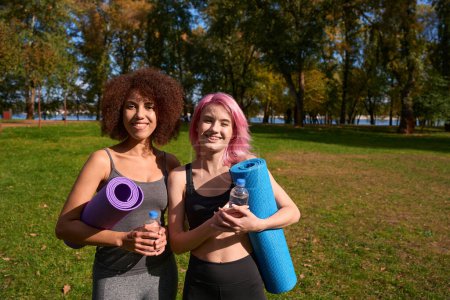 Téléchargez les photos : Deux athlètes féminines souriantes avec tapis de yoga enroulés et bouteilles d'eau debout à l'extérieur - en image libre de droit