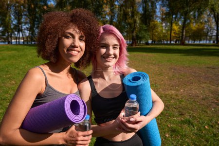 Photo for Waist-up portrait of joyous sportswomen with rolled-up exercise mats and bottles of water standing outside - Royalty Free Image