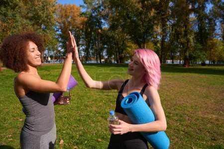 Téléchargez les photos : Athlètes féminines gaies avec des tapis d'exercice enroulés et des bouteilles d'eau se donnant cinq hauts à l'extérieur - en image libre de droit