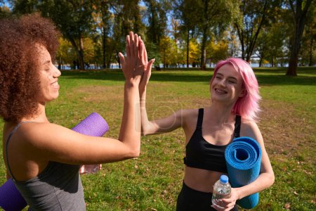 Téléchargez les photos : Joyeuses femmes athlétiques avec des tapis de yoga enroulés et des bouteilles d'eau se donnant mutuellement cinq dans le parc public - en image libre de droit