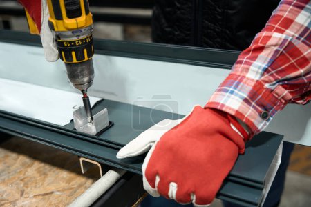 Photo for Man in protective gloves is driving screw into metal detail with electric screwdriver in workshop - Royalty Free Image