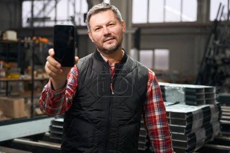 Foto de Hombre adulto mostrando el teléfono a la cámara, de pie cerca de la mesa en la producción de perfiles de acero - Imagen libre de derechos