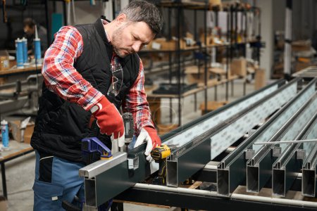 Téléchargez les photos : Homme en gants de protection assemblant une structure profilée en acier sur la table de l'atelier - en image libre de droit