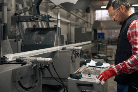 Téléchargez les photos : Homme en gants de protection travaillant à la machine de découpe de profilés métalliques dans l'usine - en image libre de droit
