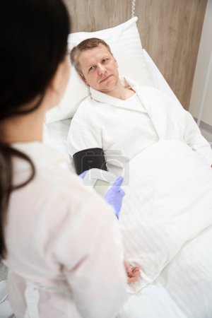 Foto de Retrato de la cintura hacia arriba del guapo paciente caucásico acostado en la cama médica mientras habla con un médico profesional en el hospital - Imagen libre de derechos