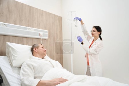 Téléchargez les photos : Portrait taille haute de gentille infirmière en vêtements blancs regarde à l'homme patient tout en préparant compte-gouttes pour lui dans la salle de maladie - en image libre de droit
