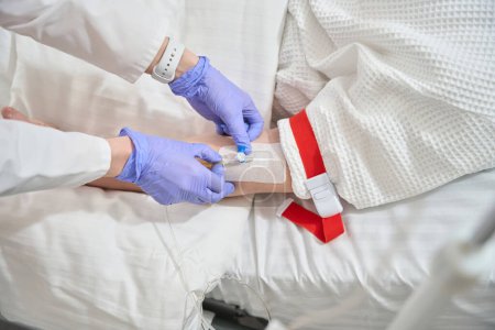 Téléchargez les photos : Image rapprochée des mains du médecin dans les gants prépare perfusion goutte à goutte pour le patient non reconnu dans la salle de maladie - en image libre de droit
